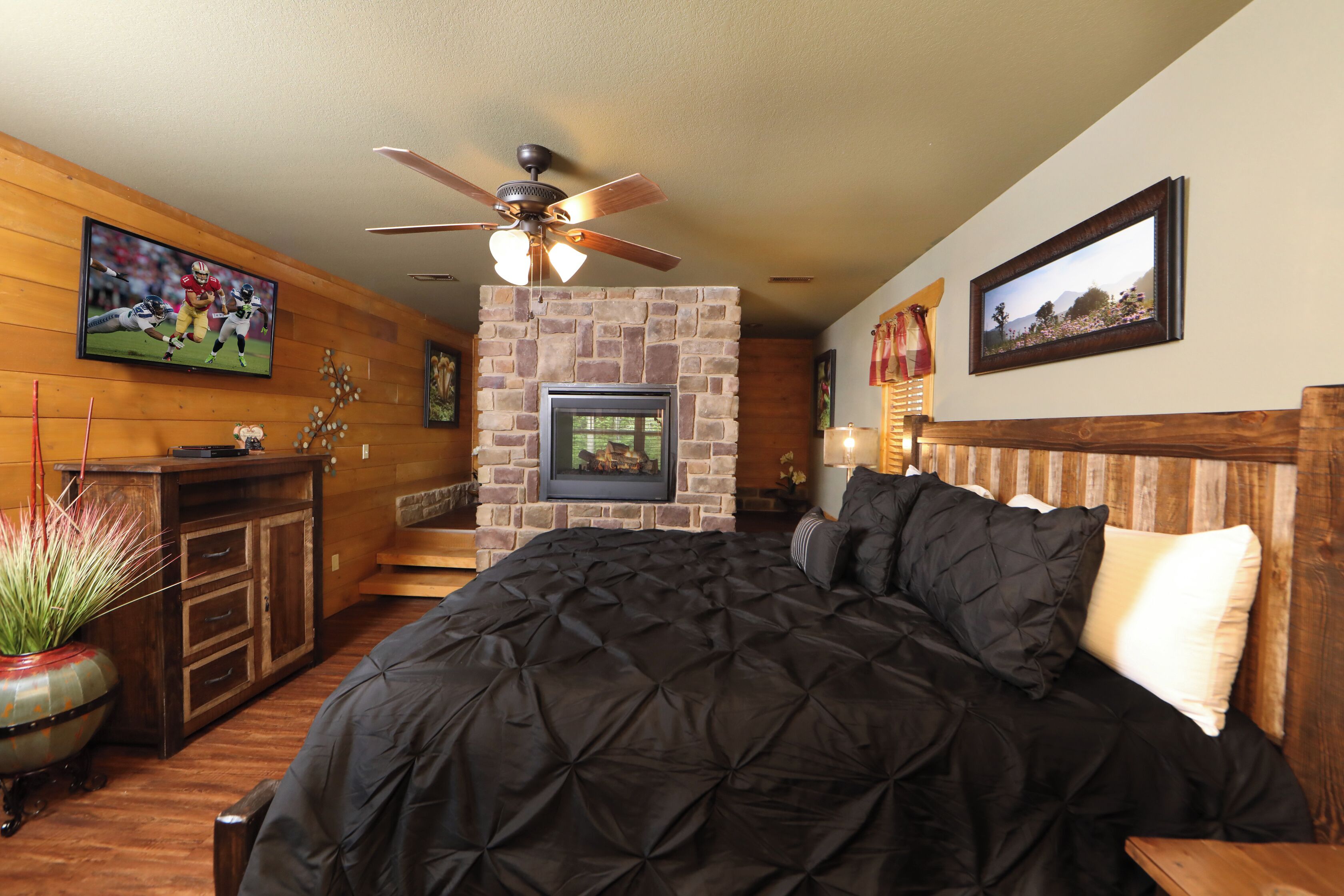 A bedroom with a large bed, a ceiling fan, a stone fireplace, wooden walls, a television, and a framed picture on the wall.