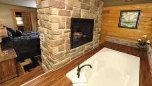 A bathroom with a stone wall, a fireplace, a large bathtub, and a wooden floor. - Head Over Hills (Gatlinburg)