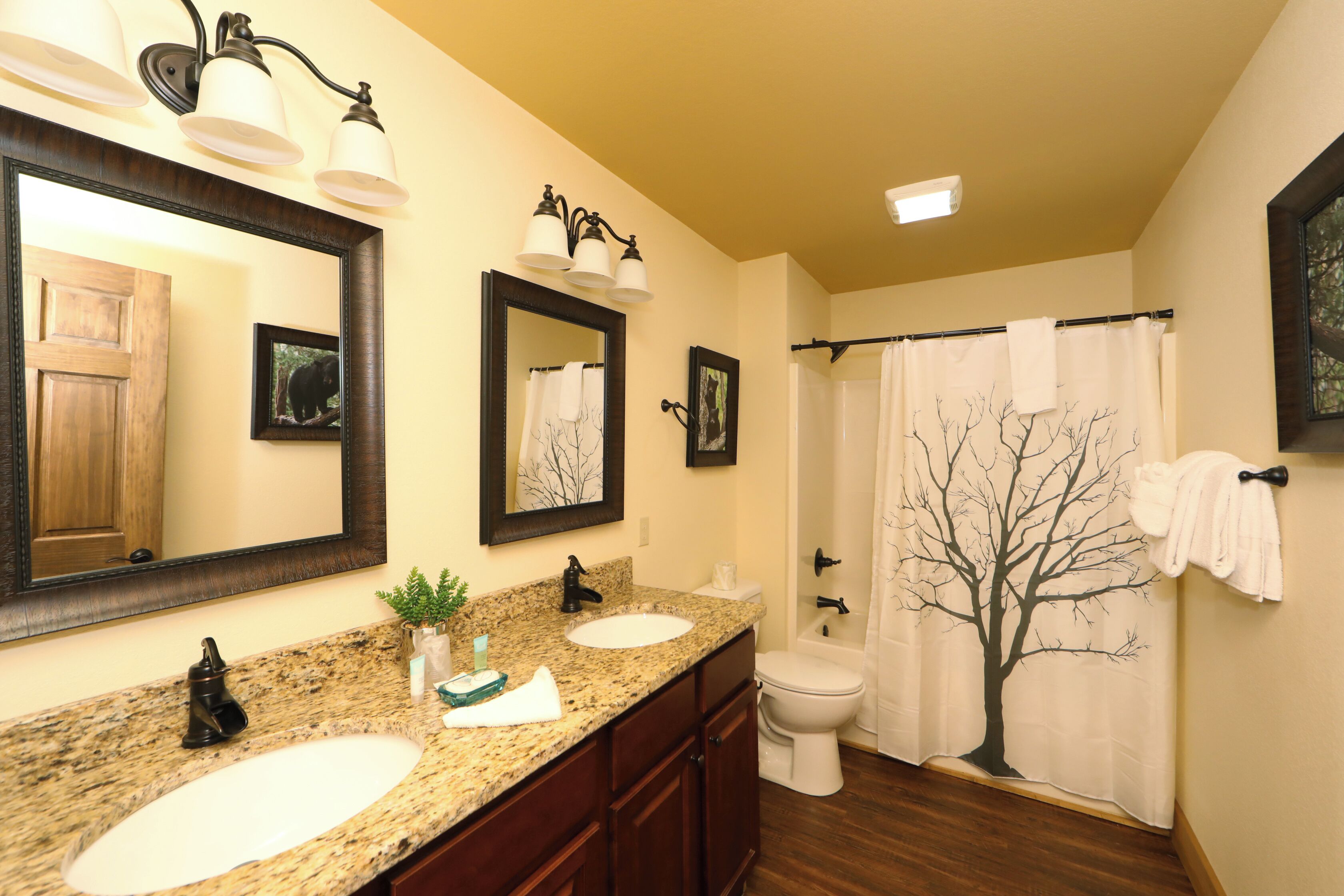 A bathroom with two sinks, a toilet, a shower with a tree-themed curtain, and a mirror with lights.