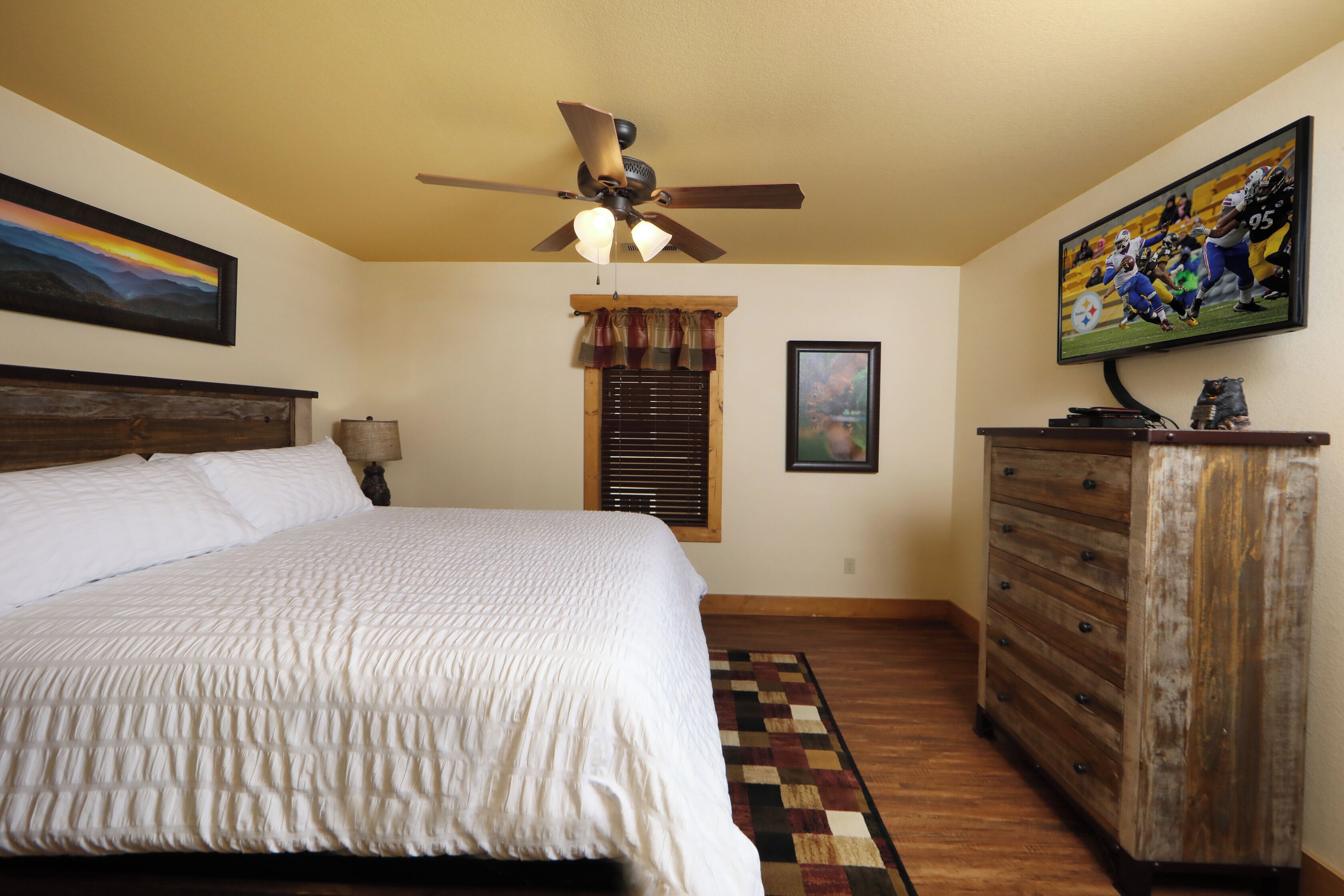 A bedroom with a bed, a ceiling fan, a television, a dresser, and a window with blinds.