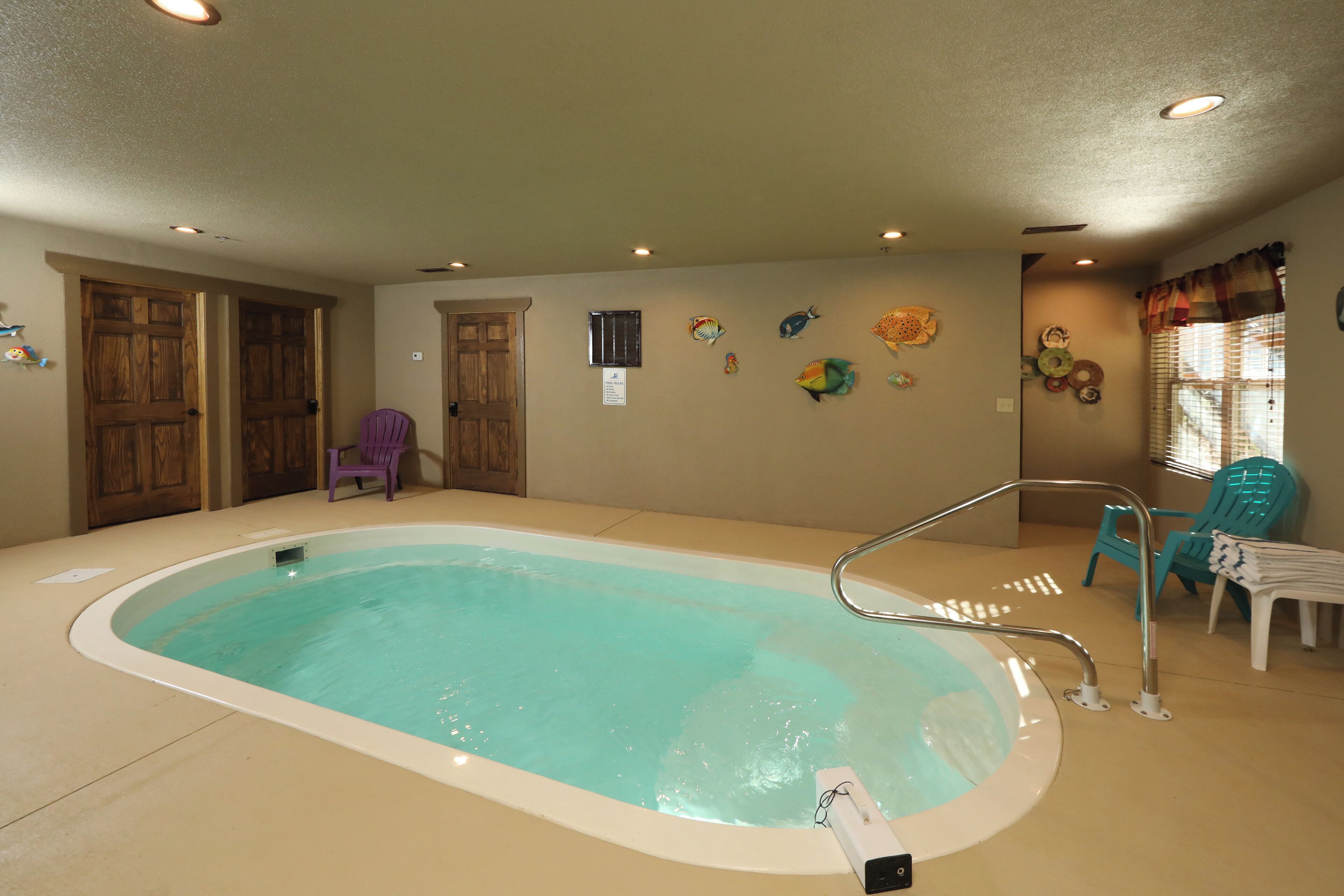 Una piscina interior con suelo de baldosas, una escalera metálica y un panel de control. Hay puertas de madera, una silla morada y una ventana con cortinas.