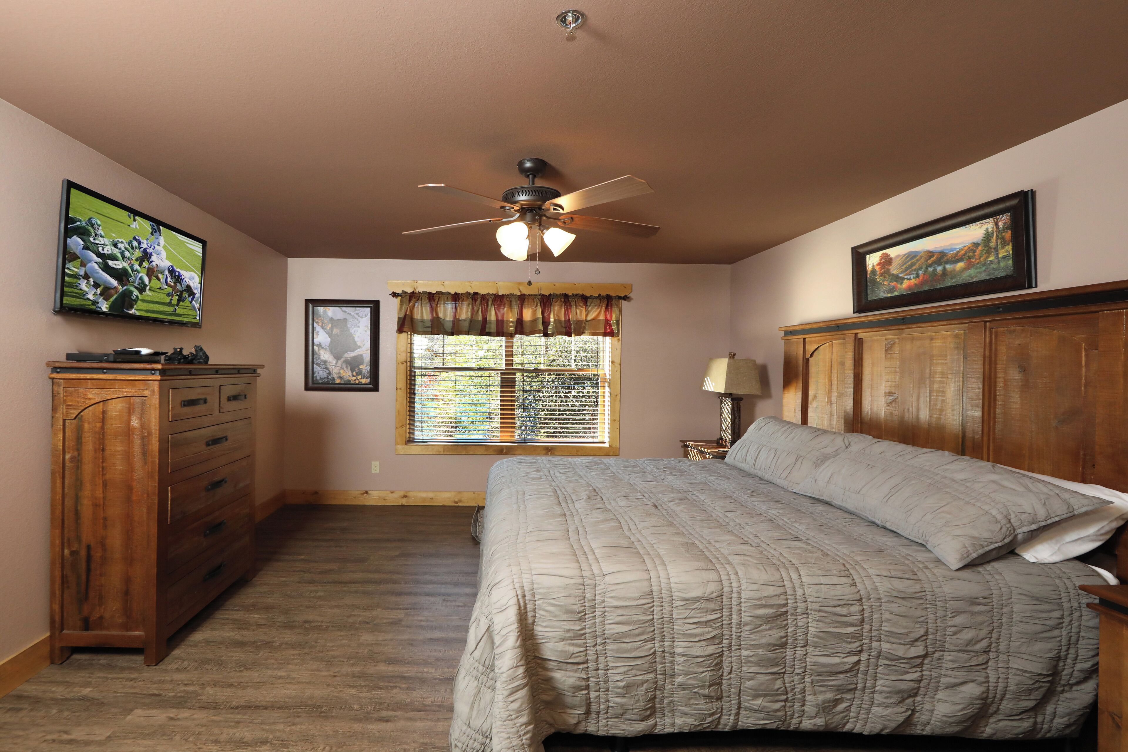 Une chambre à coucher avec un lit, un ventilateur de plafond, une télévision, une fenêtre avec des rideaux, une commode en bois et un tableau encadré accroché au mur.