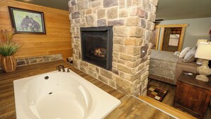 A bathroom with a stone fireplace, a large bathtub, wooden flooring, and a cozy seating area with a lamp and pillows. - Ivie Holler (Gatlinburg)