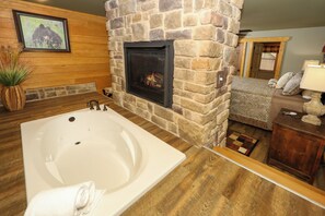 Ein Badezimmer mit Kamin aus Stein, einer großen Badewanne, Holzboden und einer gemütlichen Sitzecke mit Lampe und Kissen.