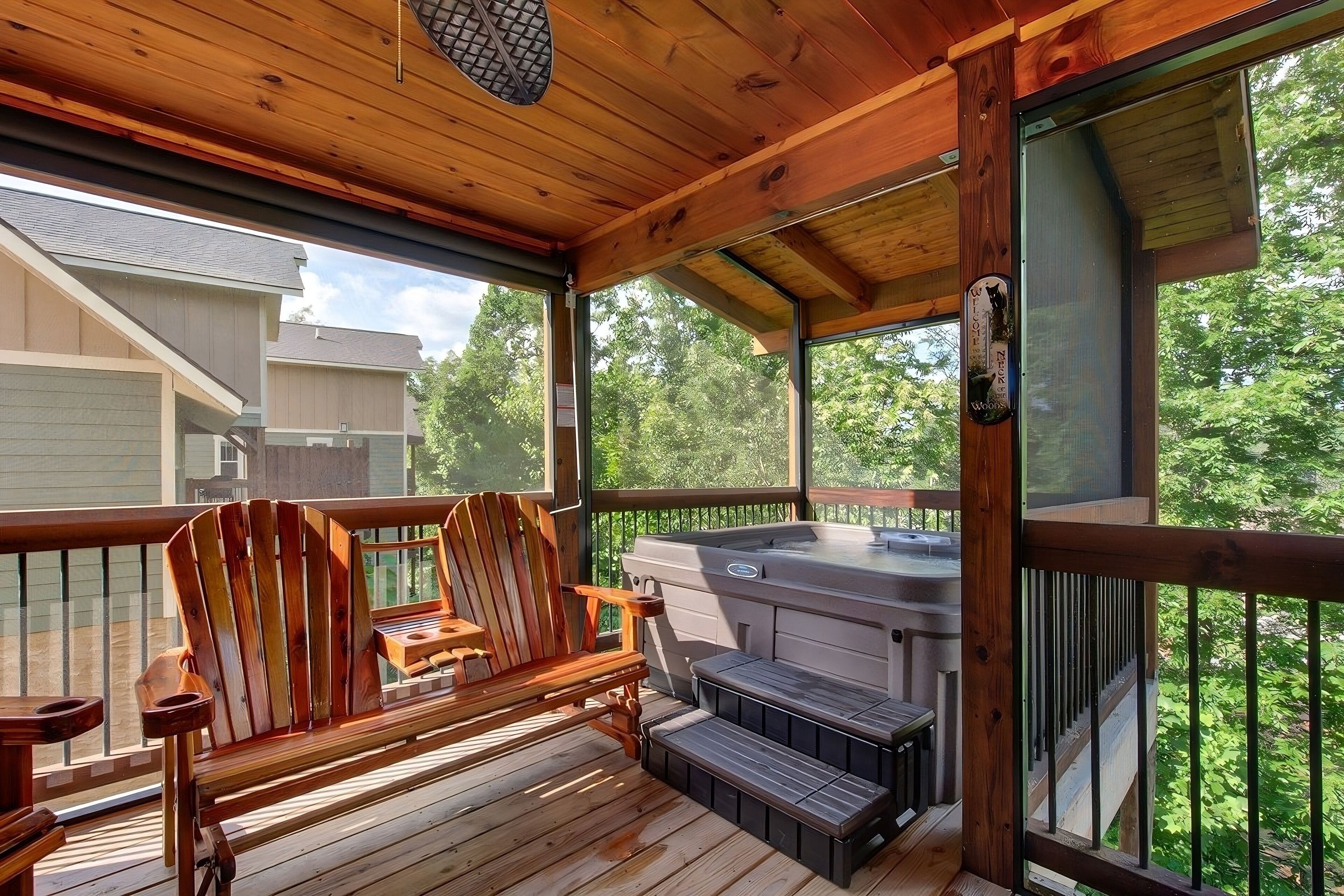 Una terraza de madera con sillas tipo Adirondack, un jacuzzi y vistas a una casa y árboles.