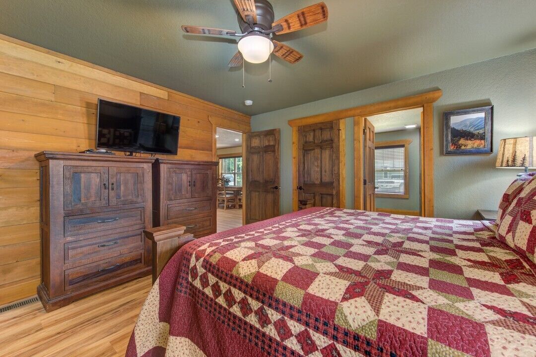 Une chambre avec une armoire en bois, un lit recouvert d’une courtepointe, un ventilateur de plafond et un téléviseur posé sur une commode en bois.