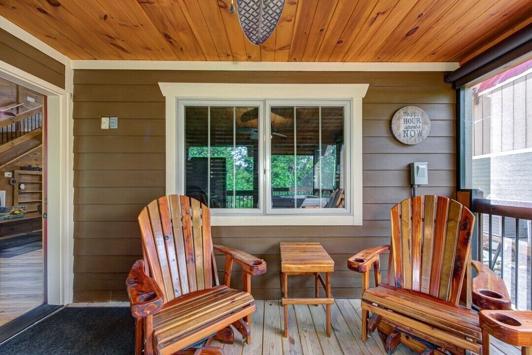 A wooden deck with two Adirondack chairs and a small table, a window with a view of greenery, and a 'Happy Hour' sign on the wall.