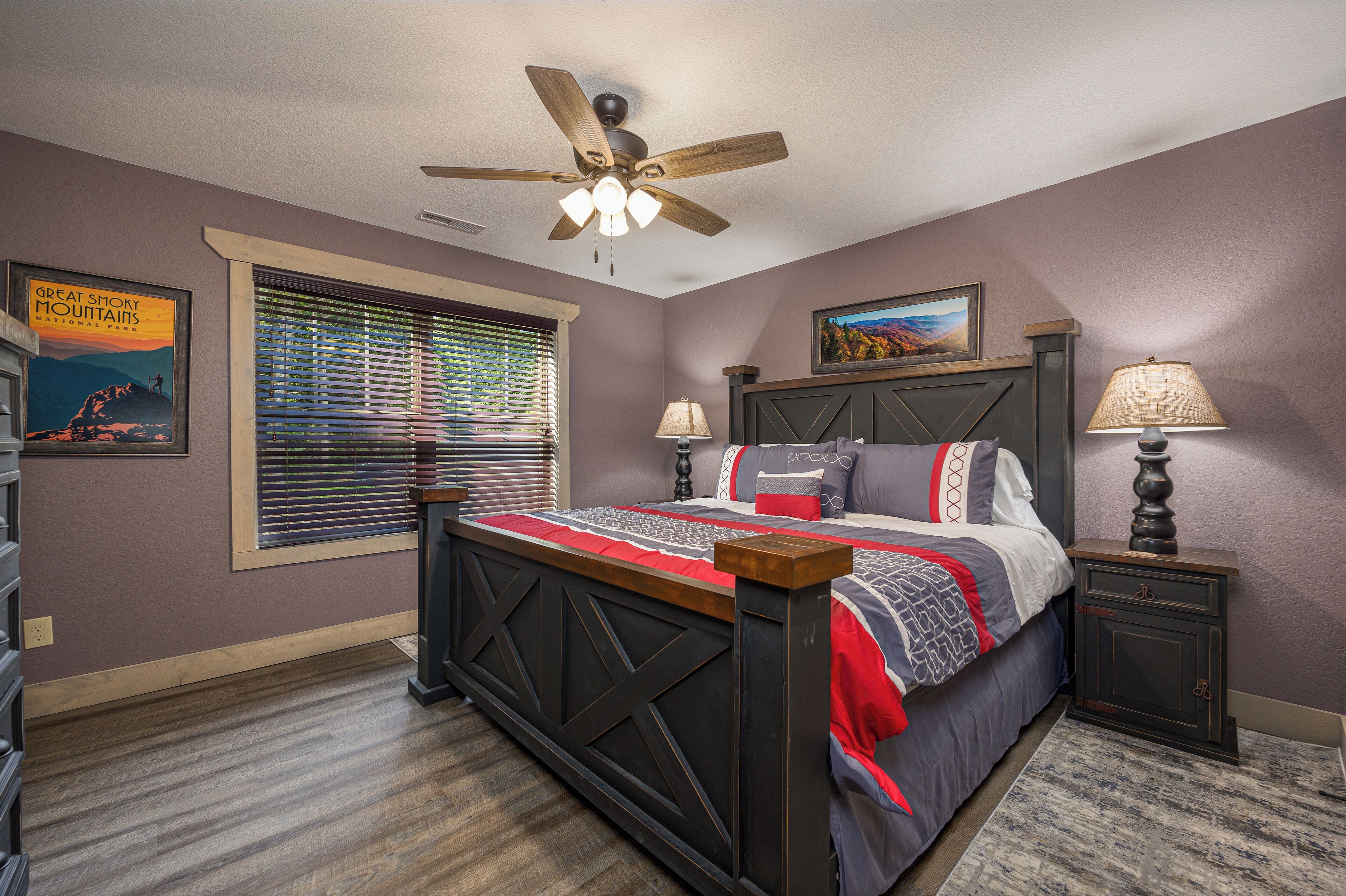 A bedroom with a bed, a ceiling fan, a nightstand, a lamp, a window with blinds, and a framed picture on the wall.
