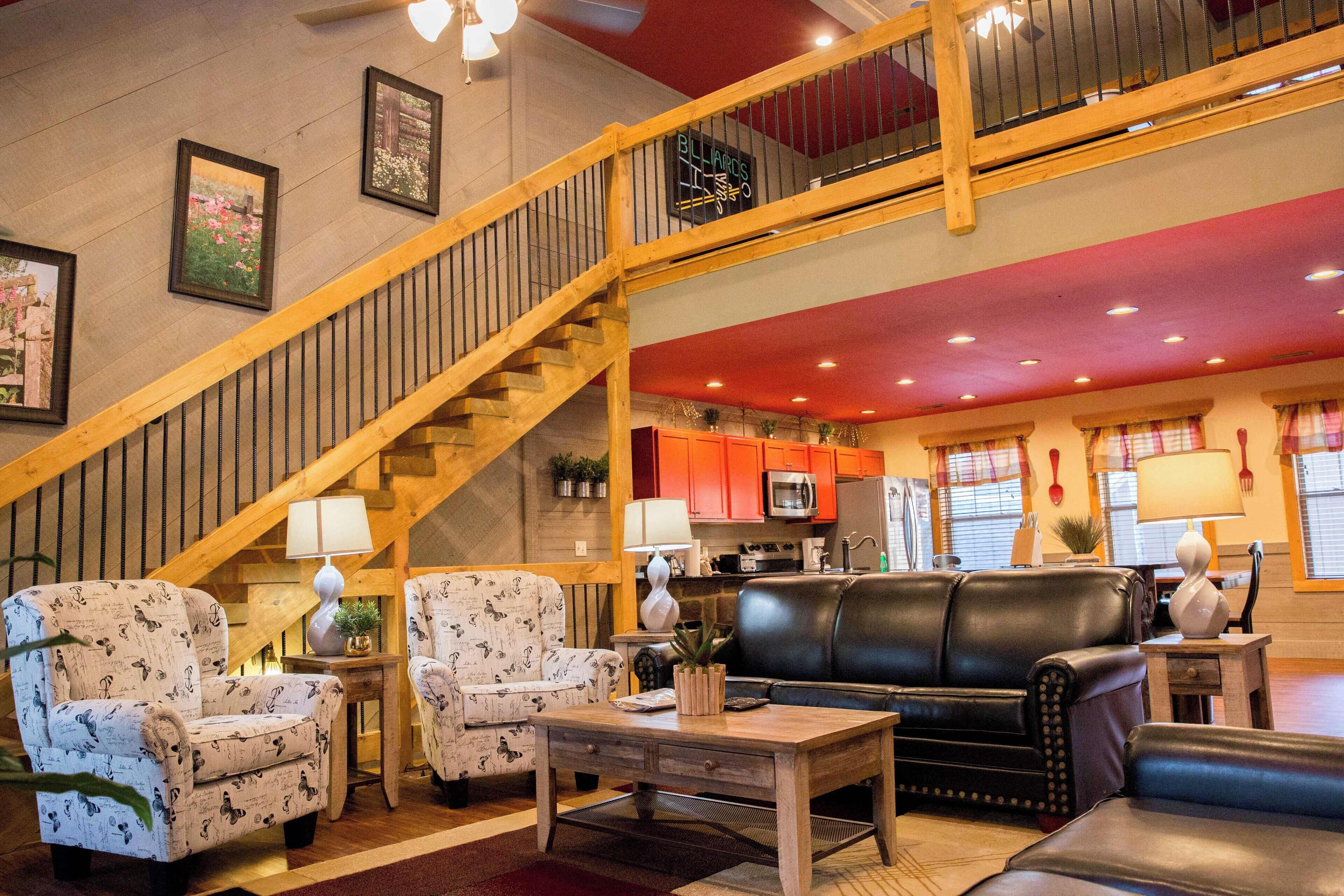 A spacious living area with a wooden staircase, a black leather sofa, patterned armchairs, and a dining table.