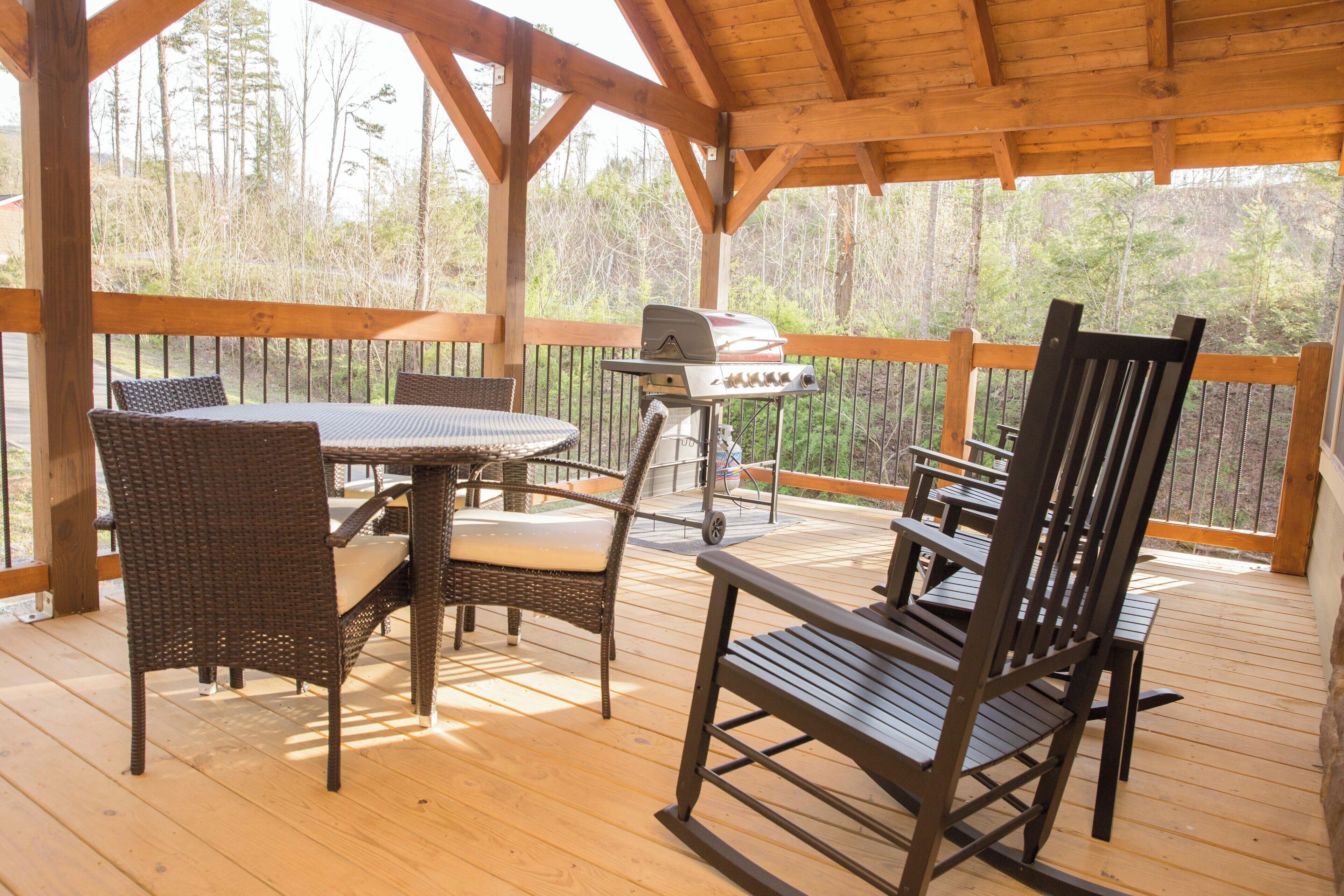 A covered outdoor deck with a table and chairs, a grill, and a view of trees.