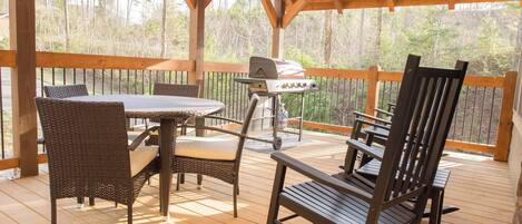 A covered outdoor deck with a table and chairs, a grill, and a view of trees.