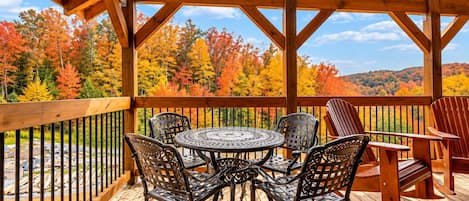 Una terraza de madera con una mesa y sillas, rodeada por una barandilla, con vistas a un bosque de follaje otoñal.