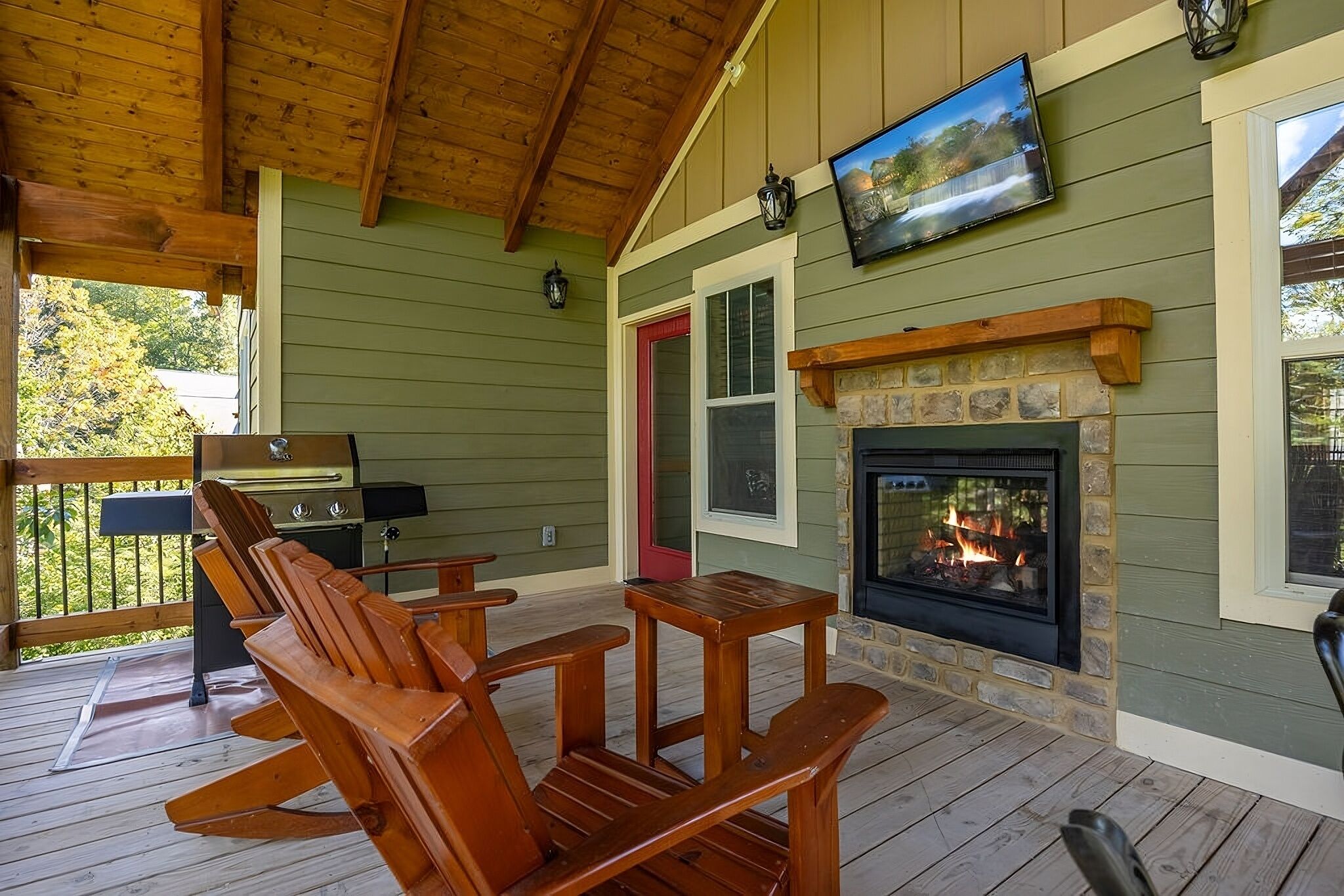 A covered outdoor seating area with wooden chairs and tables, a stone fireplace, and a television mounted above the fireplace.