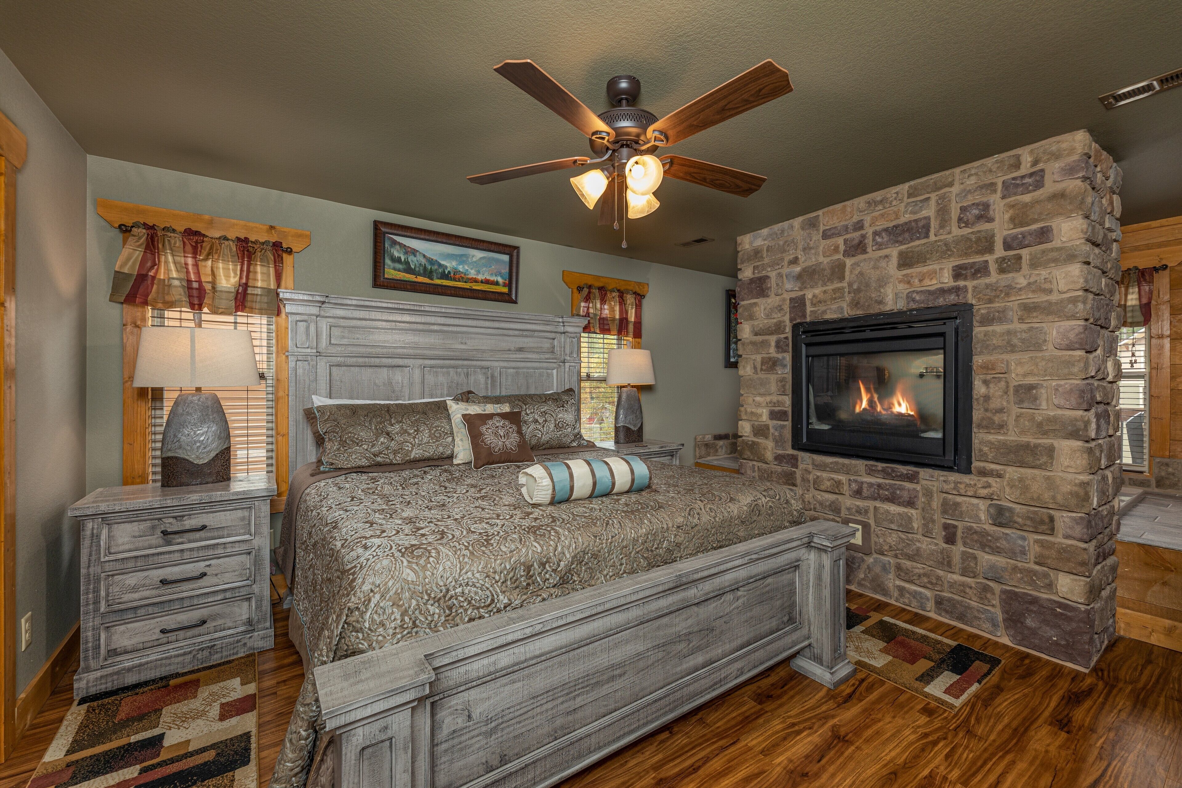 A bedroom with a stone fireplace, a large bed with patterned bedding, a ceiling fan, and a nightstand with a lamp.