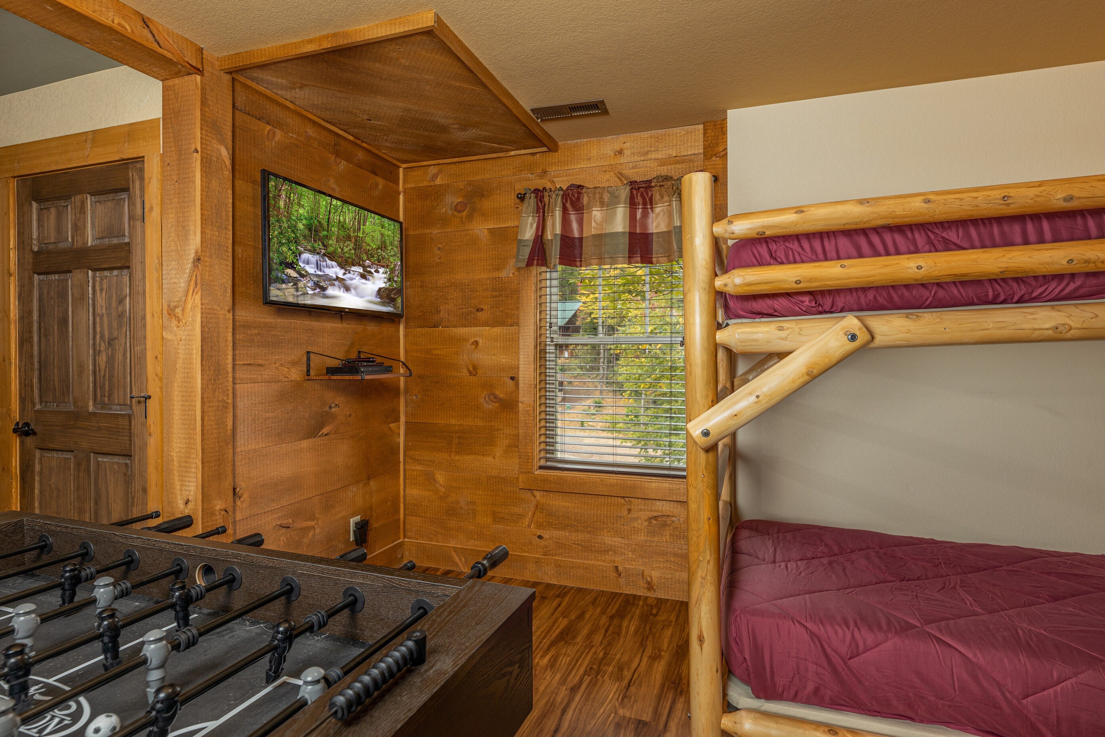 A wooden room with a foosball table, a TV mounted on the wall, and a window with curtains.