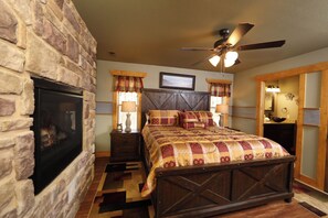 A bedroom with a stone fireplace, a bed with a quilt, a ceiling fan, and a nightstand with a lamp. - Firefly Nights (Gatlinburg)