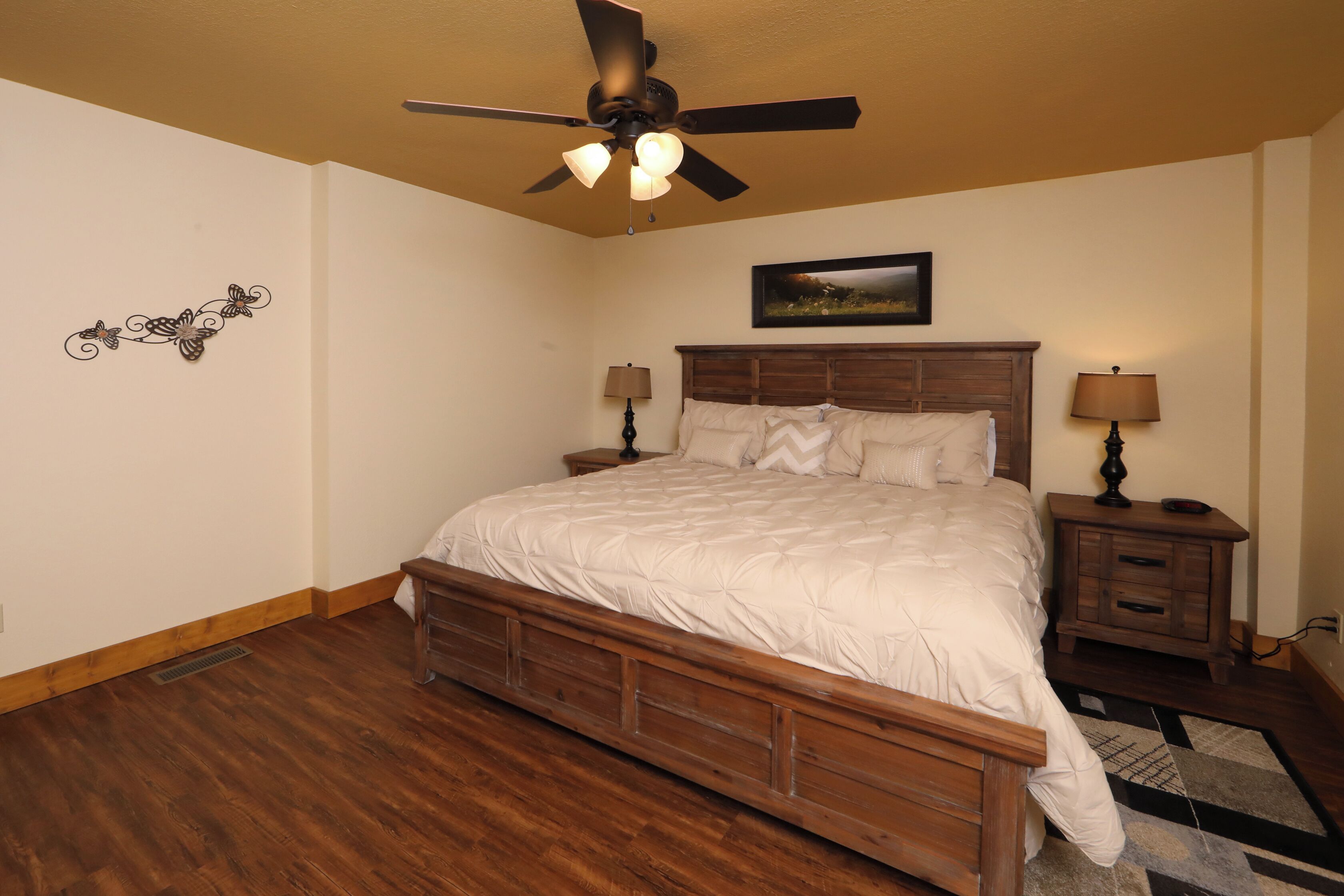 A bedroom with a wooden bed, a ceiling fan, two bedside tables with lamps, a wall-mounted artwork, and a patterned rug.