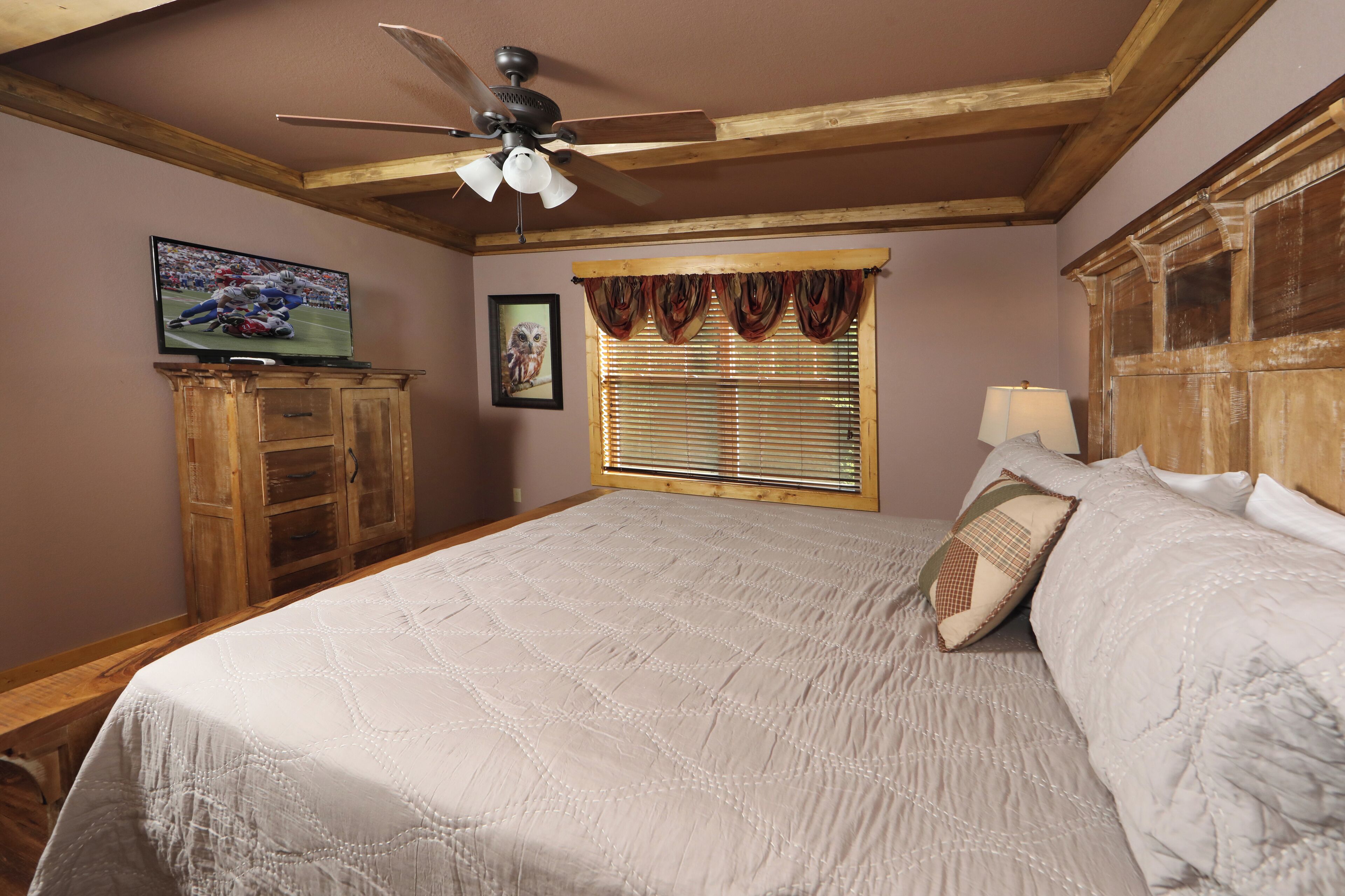 A bedroom with a large bed, wooden headboard, ceiling fan, wooden dresser, TV, window with blinds, and a framed picture on the wall.