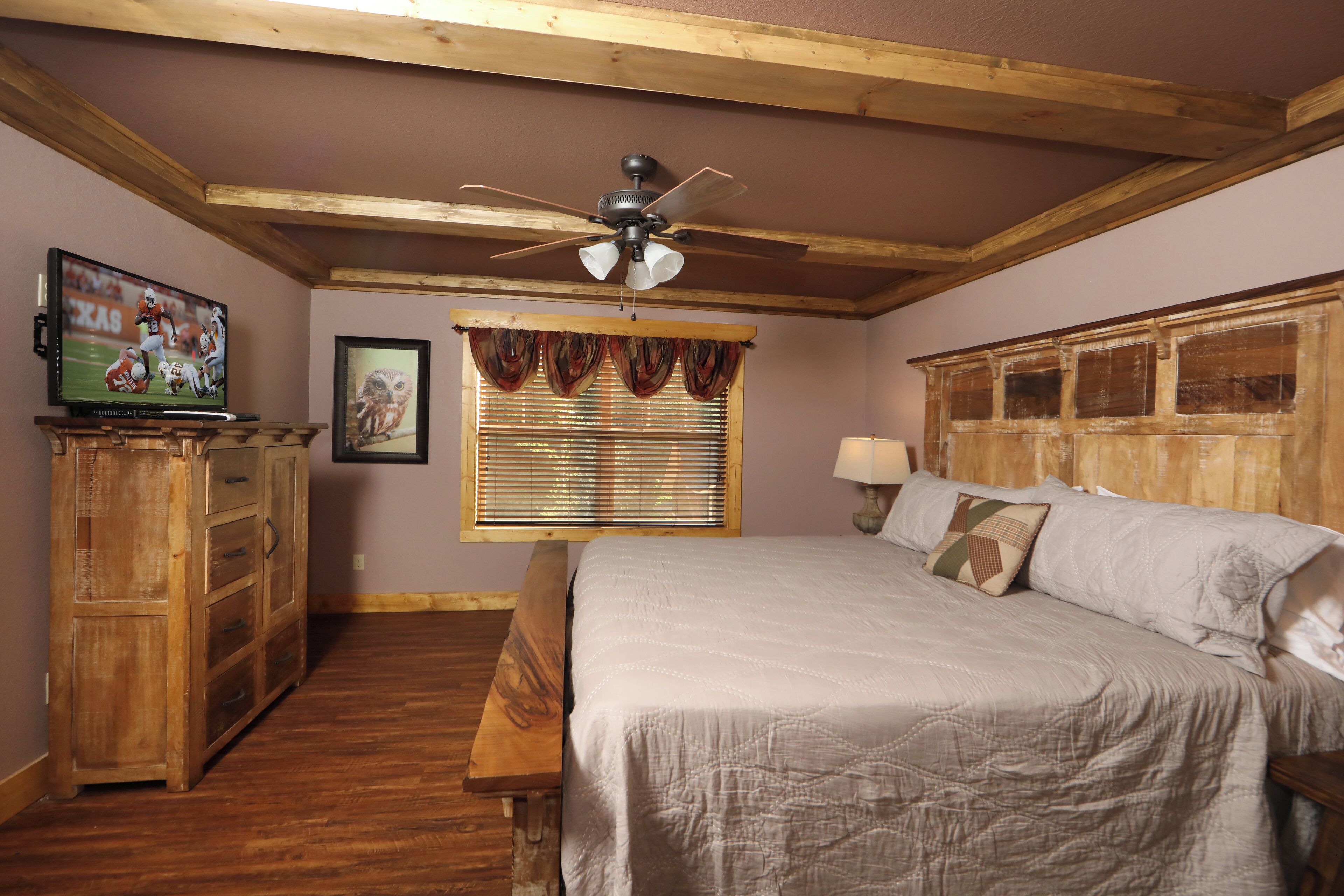 A bedroom with a wooden ceiling, a bed with a quilt, a wooden dresser, a window with blinds, and a ceiling fan.