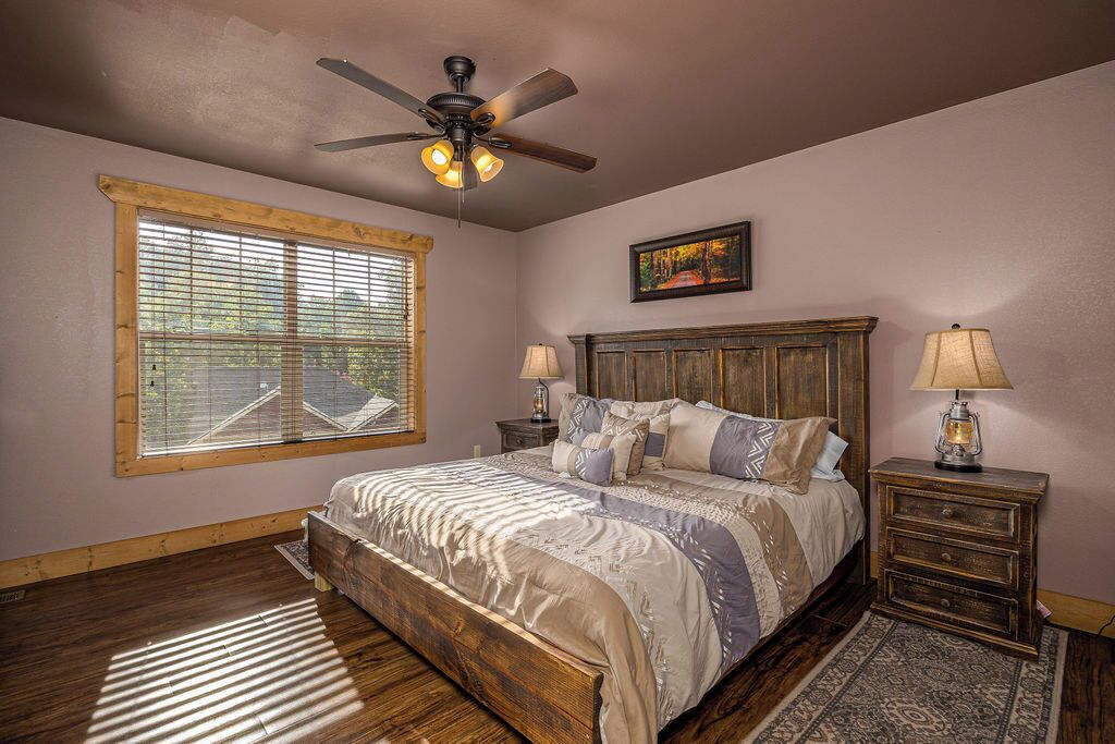 A bedroom with a bed, a ceiling fan, a window with blinds, a nightstand with a lamp, and a rug.