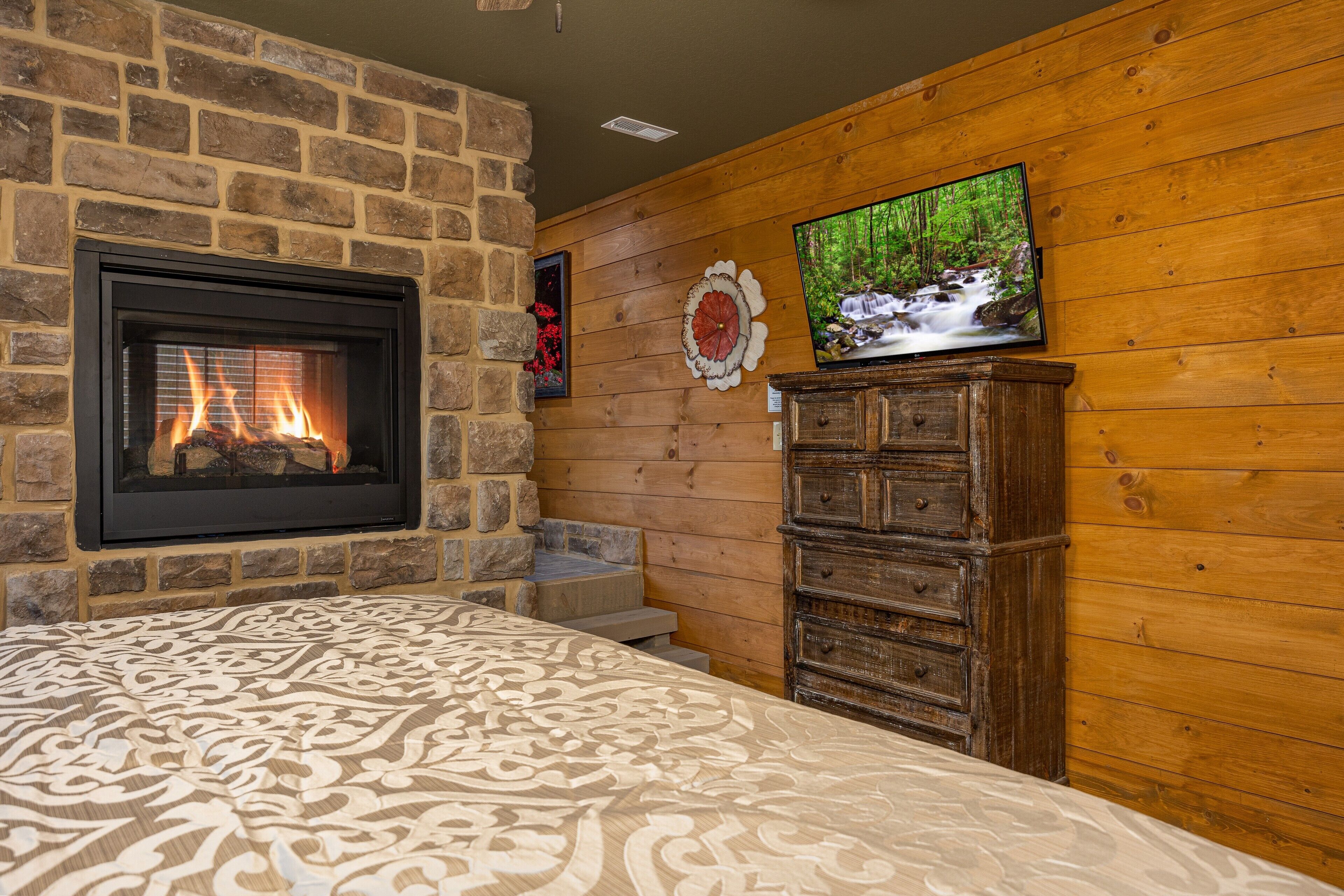 A room with a stone fireplace, a television mounted on a wooden wall, and a wooden dresser.