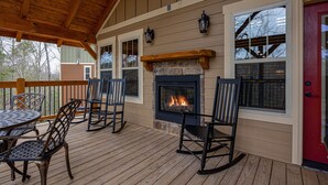 A covered deck with a stone fireplace, wooden railing, and metal chairs. - Serenity Peak (Gatlinburg)