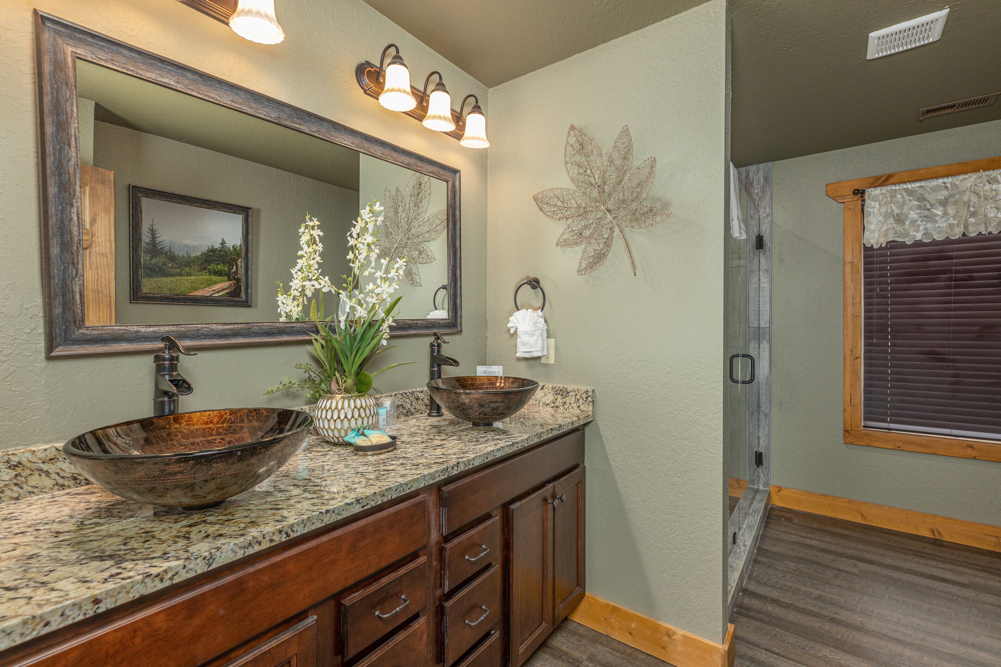 A bathroom with a large mirror, two sinks with a vessel sink, wooden cabinets, and a decorative leaf on the wall.