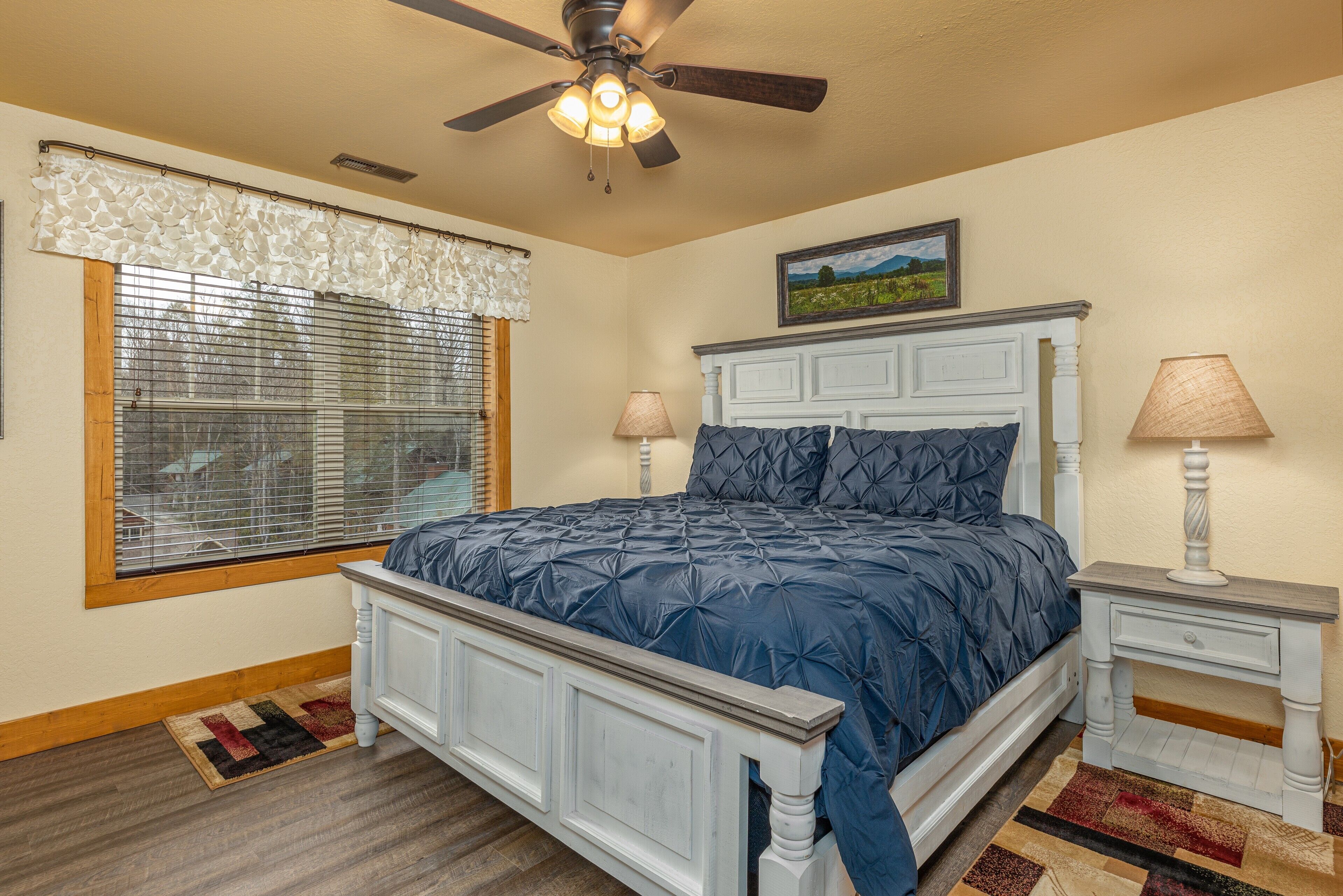A bedroom with a bed, two bedside tables, a ceiling fan, a window with blinds, and a rug.