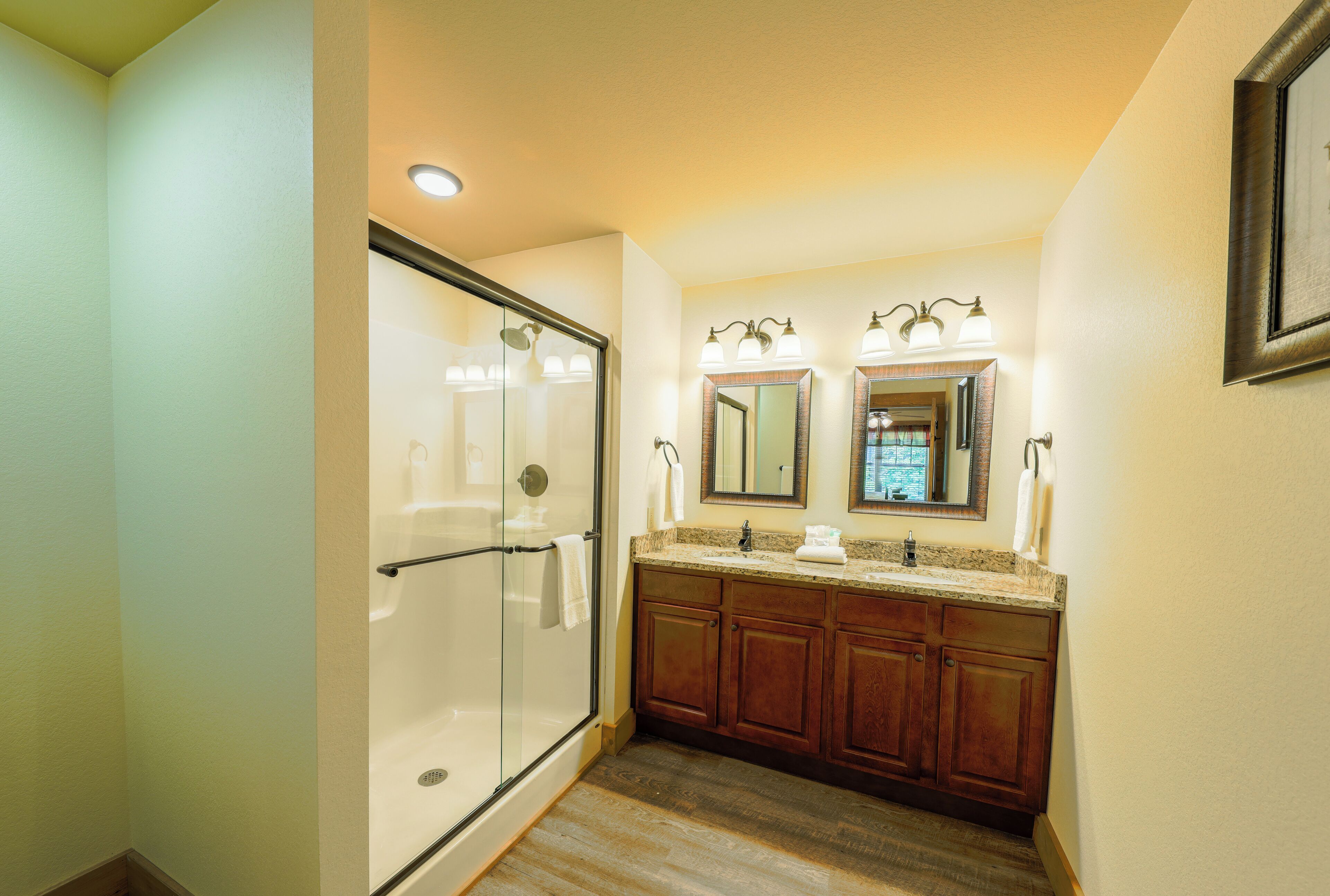 A bathroom with a glass shower enclosure, wooden cabinets, a double sink with a granite countertop, and wall-mounted lighting.