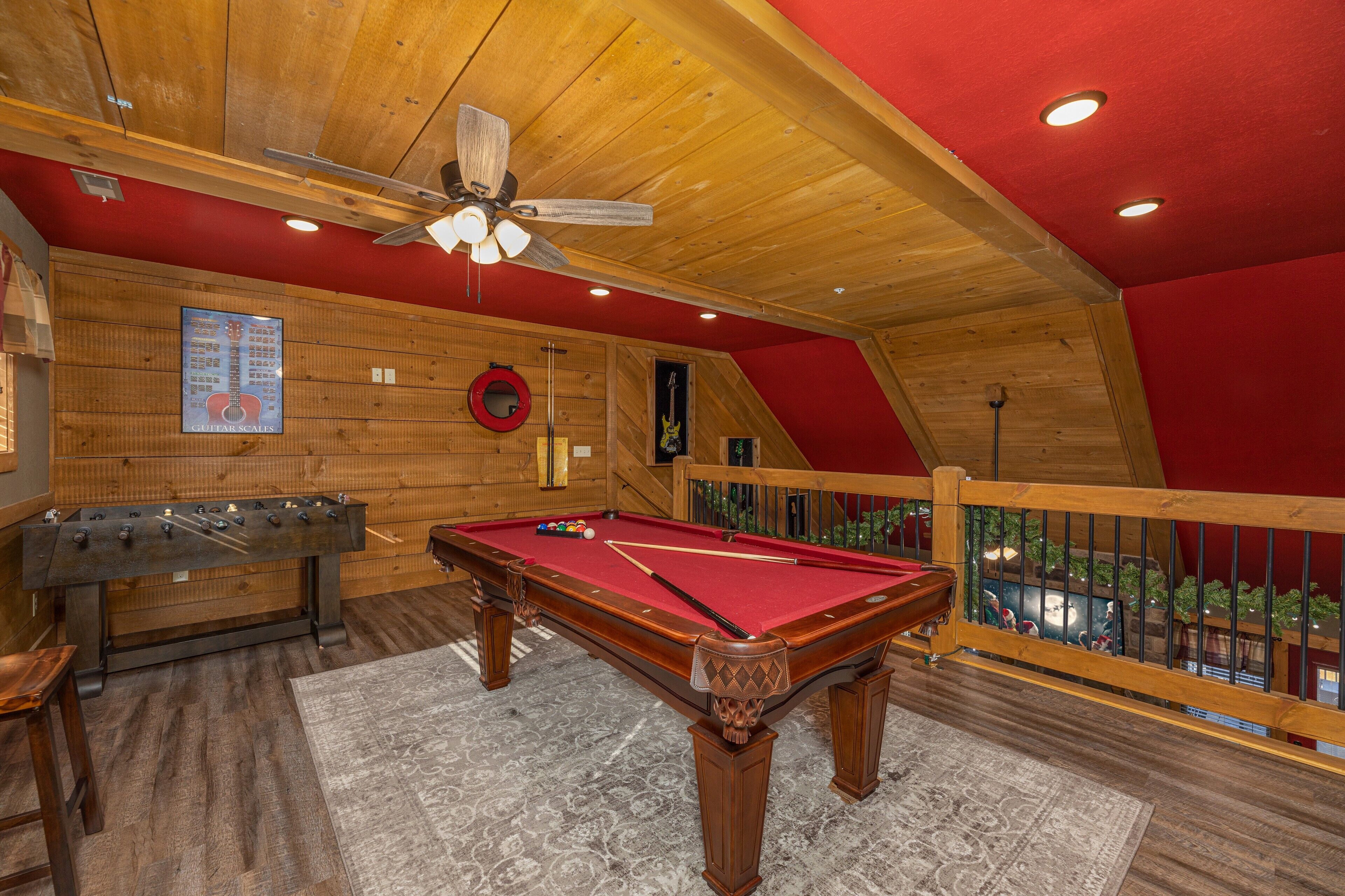 Une table de billard recouverte d’un drap de feutre rouge, une table de baby-foot et un ventilateur de plafond dans une pièce aux murs en bois et dont un des murs est peint en rouge.
