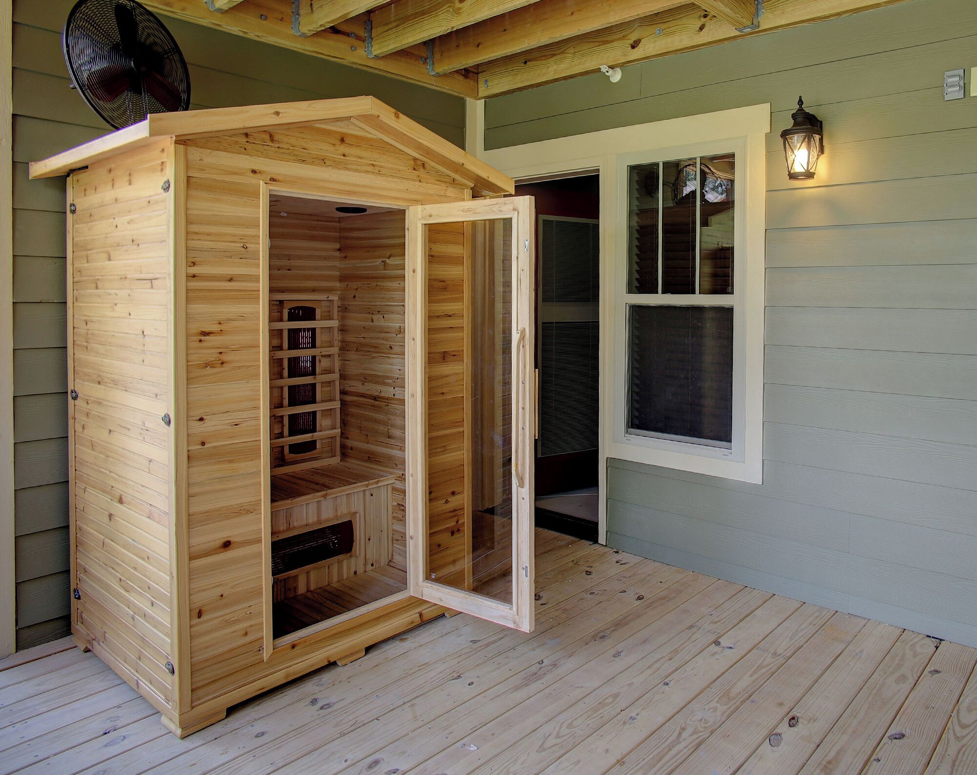 A wooden sauna structure with a sliding door, a fan, and a window with a lantern.