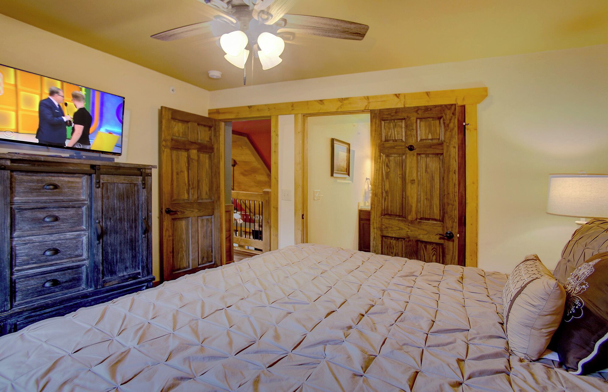 A bedroom with a large bed, wooden doors, a ceiling fan, and a television on a stand.