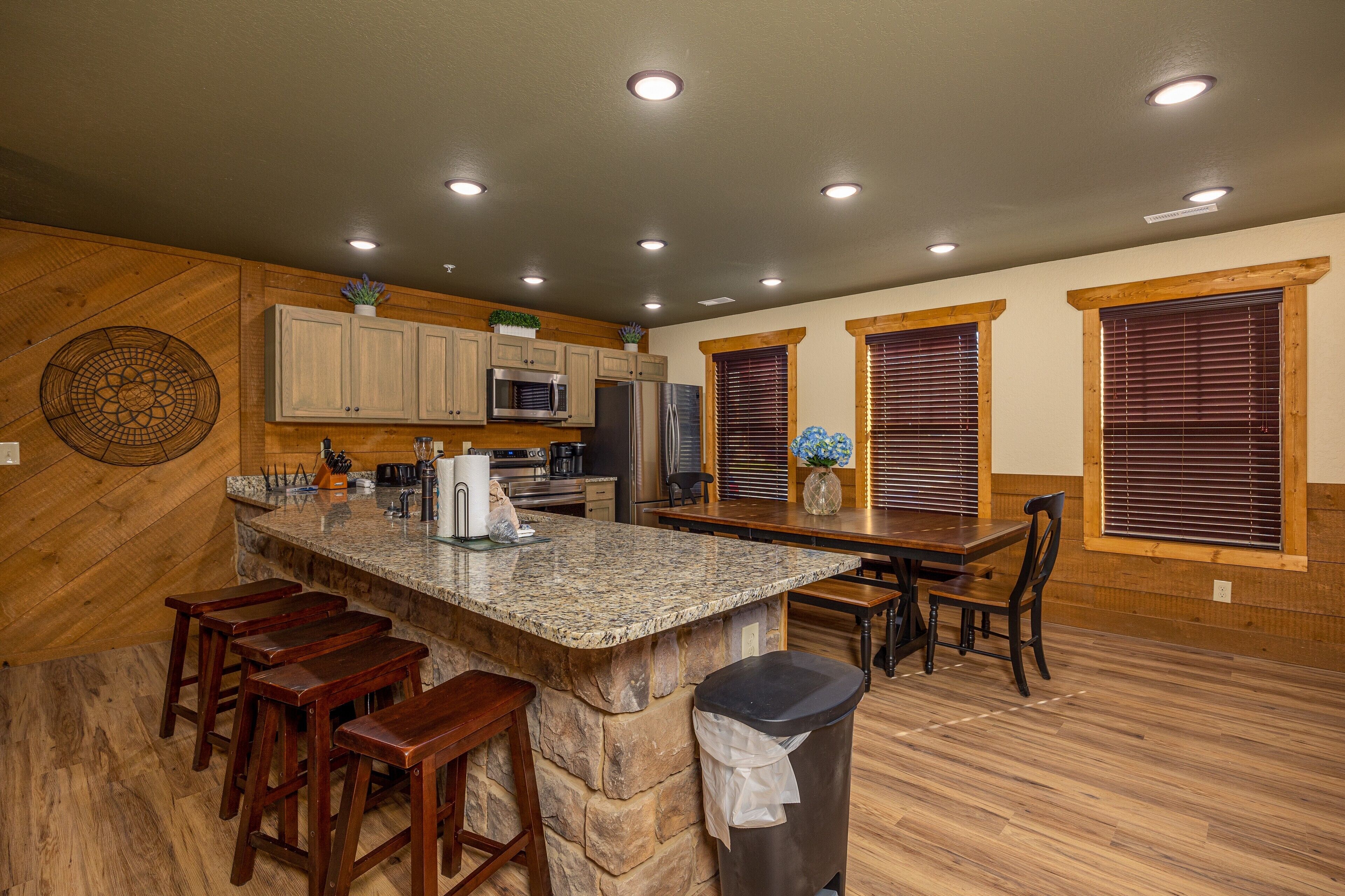 A kitchen with a granite island, wooden bar stools, a trash can, and wooden flooring.