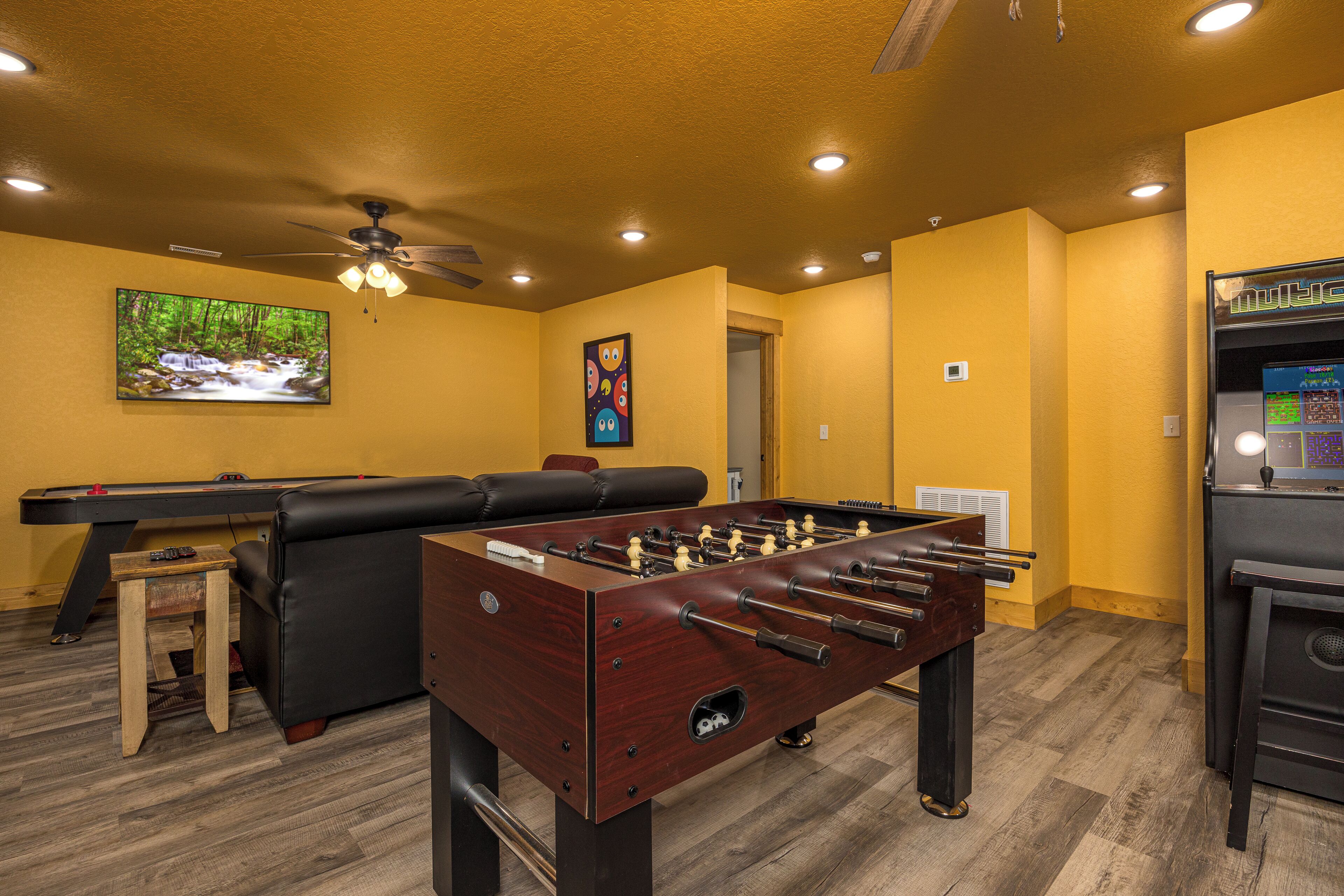 A room with a foosball table, a pool table, a sofa, and a television displaying a nature scene.