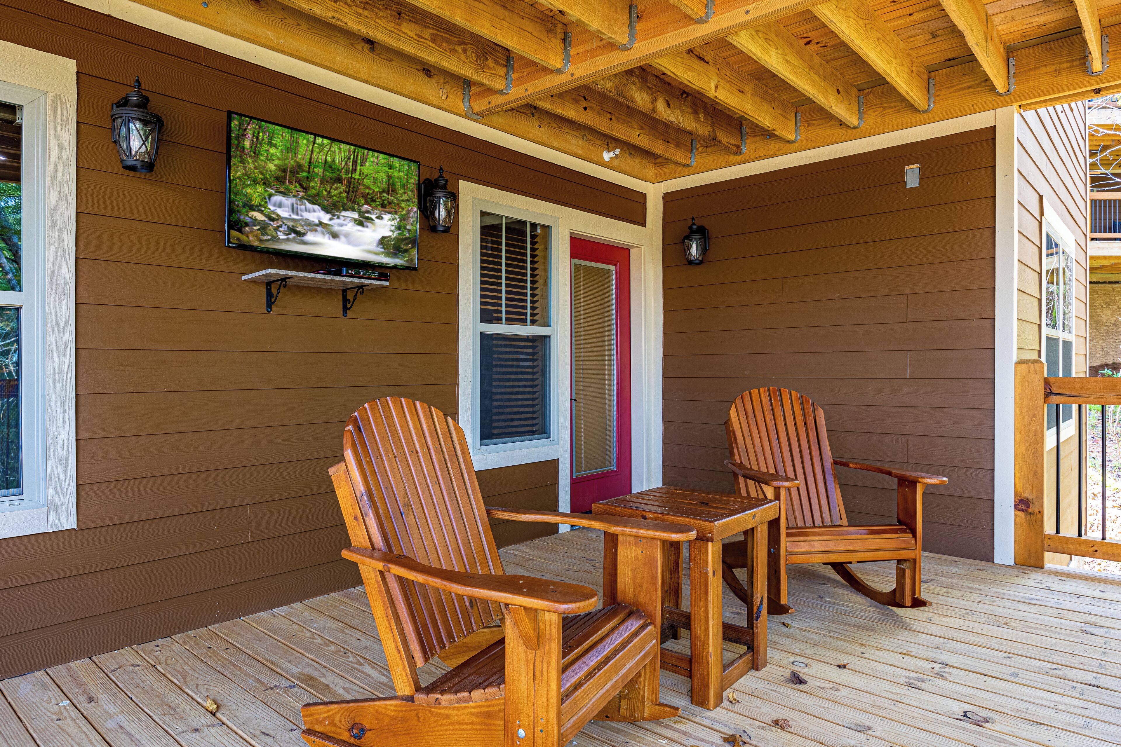 A wooden deck with two Adirondack chairs and a table, a TV mounted on the wall, and a red door.
