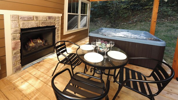 A wooden deck with a stone fireplace, a hot tub, and a round table with chairs. - Misty Mountainside (Gatlinburg)
