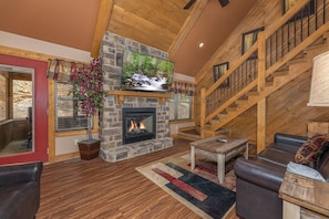 A cozy living room with a stone fireplace, a flat-screen TV mounted above it, a leather sofa, and a wooden staircase. - Heavenly Haven (Gatlinburg)