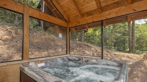 Een jacuzzi met een houten terras en grote ramen met uitzicht op een bebost heuvelgebied.