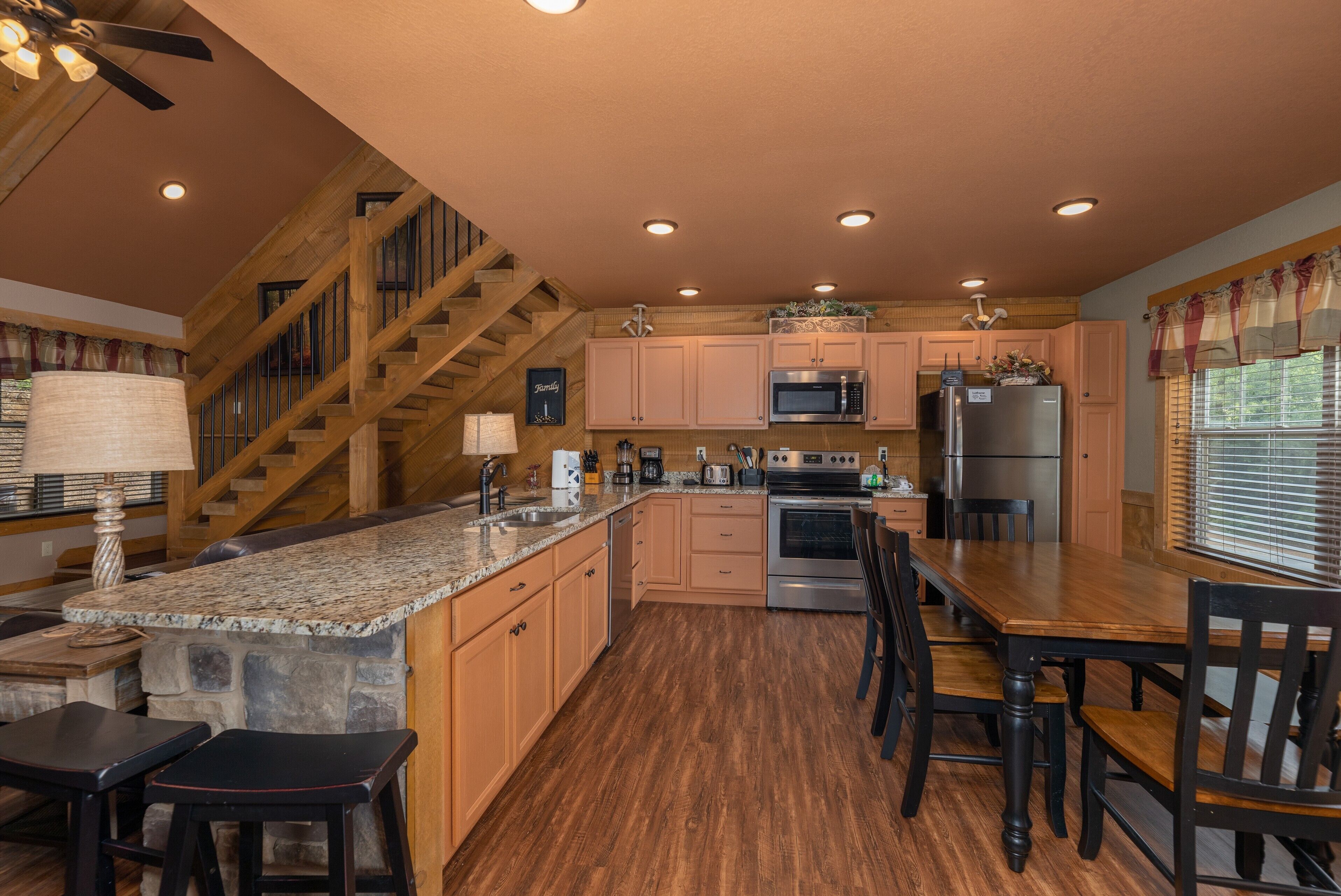 A spacious kitchen with granite countertops, wooden cabinets, and a central island. A wooden staircase leads to an upper level.