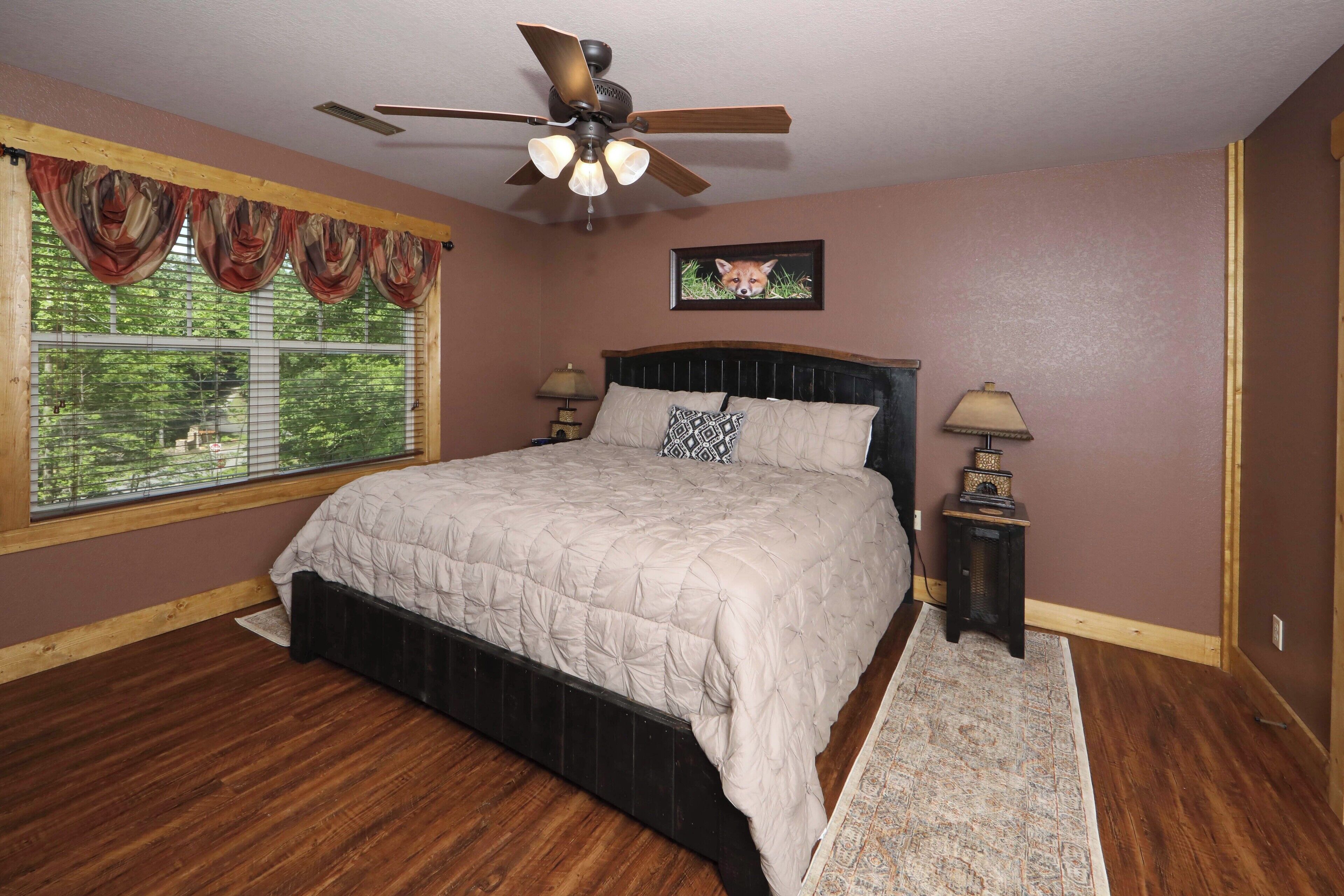 Quarto com cama grande, ventilador de teto, janela com cortinas, mesa de cabeceira com abajur e quadro na parede.