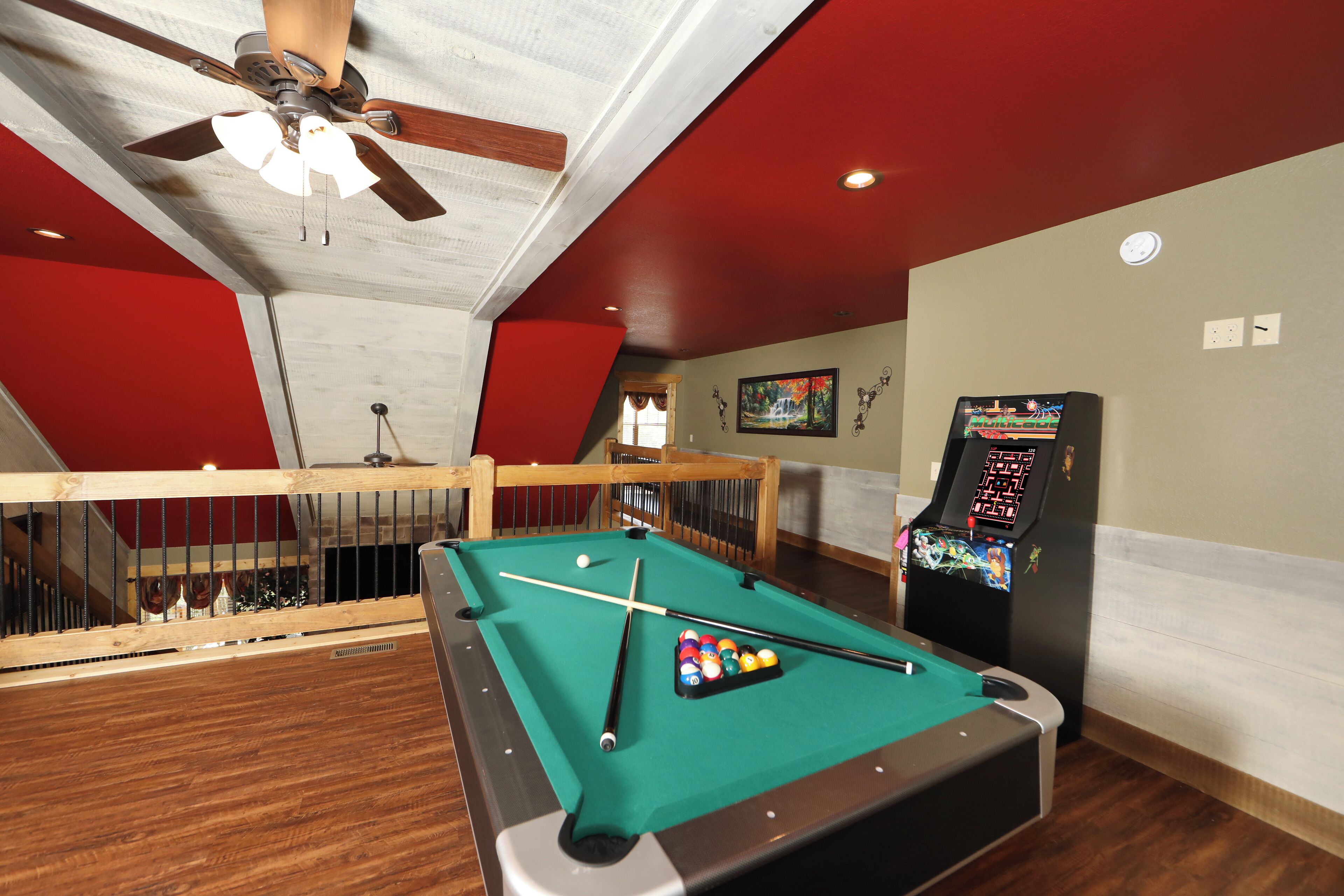 A pool table with a pool cue and balls, a ceiling fan, and a video game machine in a room with red and white walls.