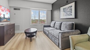 Ein modernes Wohnzimmer mit einem grauen Sofa, einem kleinen runden Tisch, einem Fernseher links und einem Fenster mit Jalousien.