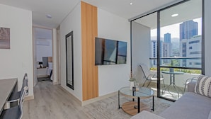 Una sala moderna con sofá, una mesa de centro de vidrio, televisor de pantalla plana montado en la pared y un balcón con vistas a la ciudad.