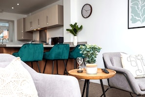 A modern living room with a sofa, two teal chairs, a small wooden table with a candle and an hourglass, a grey armchair, and a kitchen in the background.