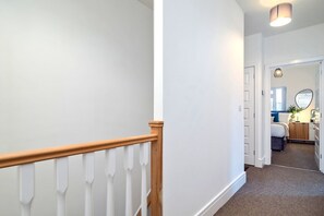 Ein Flur mit Holzhandlauf, weißen Wänden und Teppichboden, der zu einem Schlafzimmer mit Bett, Nachttisch und Spiegel führt.