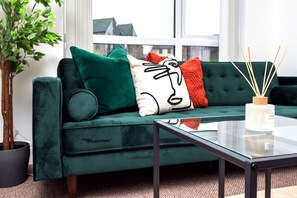 Ein grünes Sofa mit Kissen, das ein Cartoon-Gesicht zeigt, ein Glastisch und eine Pflanze in einem schwarzen Topf.