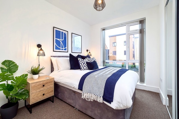 A modern bedroom with a bed, bedside tables, a potted plant, and a window with a view of buildings.