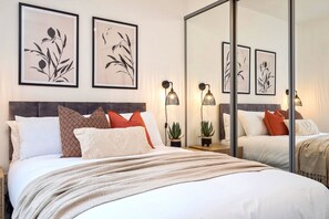 A modern bedroom with a large bed, multiple pillows, a bedside table with a lamp, and framed artwork on the wall.