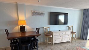 A dining area with a wooden table, black chairs, a white dresser, and a TV mounted on the wall. - Spacious Ocean Front Studio for 4 with Picture-Perfect View! (Daytona Beach Shores)