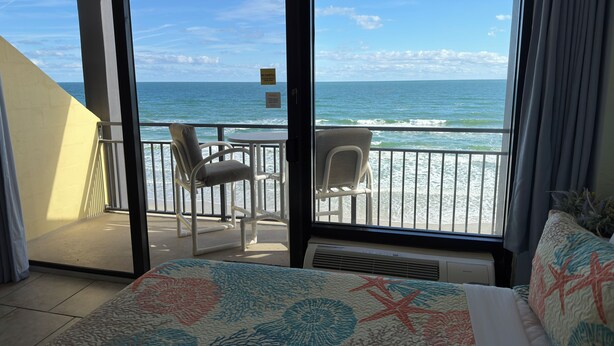 A balcony with a view of the ocean, a bed with a floral bedspread, and two chairs. - Spacious Ocean Front Studio for 4 with Picture-Perfect View! (Daytona Beach Shores)